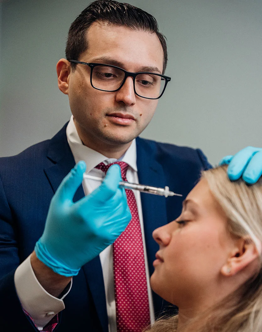 Dr. Kachare, a light-skinned man in a dark suit, glasses, and a reddish-pink patterned tie, administers an injection to a light-skinned woman with her head tilted back and eyes closed. He wears teal medical gloves, and the background is a light gray-blue wall. - Botox Wrinkle Relaxers in Greenville, NC