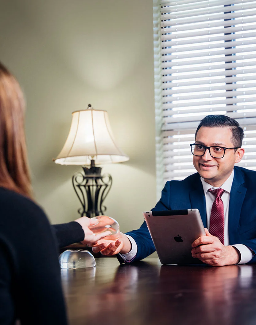 A medium shot of a consultation. A woman, seen from behind wearing a dark blouse, sits across a dark wooden table from Dr. Kachare in a navy suit and maroon tie. Dr. Kachare, wearing glasses, clasps the woman’s hands, showing a supportive connection. A beige lamp stands behind the woman, with Venetian blinds and a light-colored wall in the background. - Breast Augmentation in Greenville, NC