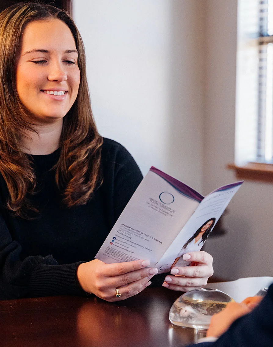 A light-skinned woman with shoulder-length brown hair sits at a dark wooden table, smiling gently while looking down at an open brochure she holds with both hands. The brochure features a circular design and text about procedures or services. Another person’s hand and arm are partially visible in the lower right, indicating a consultation or meeting. A small clear round object rests on the table between her hands. The background shows a light beige-white wall and part of a window, suggesting a medical or aesthetic office setting. - Breast Implant Exchange in Greenville, NC