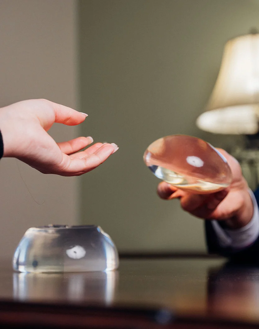 A person's hand extends toward a clear, round object, resembling a breast implant or similar medical device, resting on a dark wooden table. Another person, seated, is holding a similar object. The table surface is dark brown or mahogany, reflecting light. The background is a muted, light brownish-gray wall, with a portion of a lamp visible in the upper right quadrant. The image appears to be of a consultation or demonstration involving breast augmentation or a similar procedure. - Breast Implant Revision in Greenville, NC