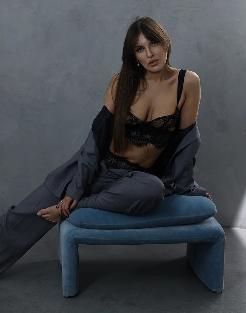 A woman with long dark brown hair sits on a modern teal bench with legs bent and feet resting together. She wears a black lace bustier top and matching wide-leg pants. Her expression is neutral, and a small light earring is visible. The backdrop is a textured gray wall, and soft natural light casts gentle shadows. - Breast Reduction in Greenville, NC