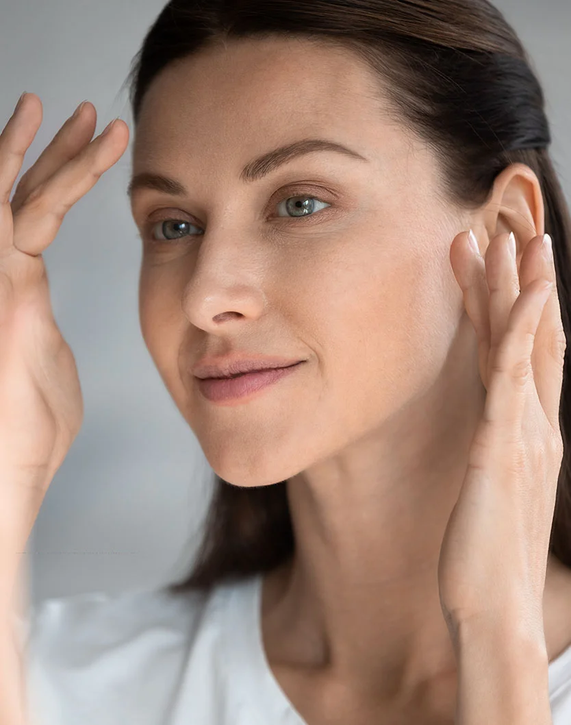 Close-up of a woman examining her face. Her hands are positioned near her forehead and cheek area, touching and feeling the skin. The woman has light-brown hair pulled back and a light complexion. She appears to be examining her skin's texture and condition. Her expression is neutral. A light-colored, likely white, top is visible. The background is a light gray or grayish-white. - Brow Lift in Greenville, NC