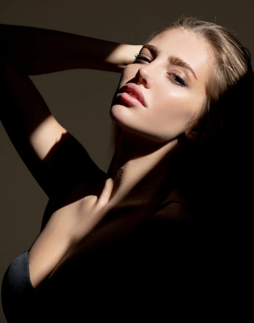 Close-up of a young woman with light brown hair, arms draped over her head, wearing a black form-fitting top. Light and shadow dramatically highlight her smooth skin and defined makeup. Her expression is relaxed and engaging, set against a dark, neutral background. - Buccal Fat Removal in Greenville, NC