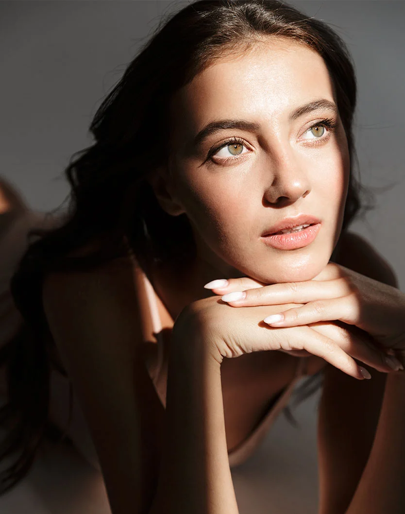 Close-up portrait of a woman with long, dark brown hair lying down, her head resting gently on her hands. She has smooth, light-toned skin, light-colored eyes, and subtle, natural makeup. Her hands are delicately positioned, with nails painted a light, nearly white shade and shaped to a slight point. Soft, dramatic shadows from above create depth and highlight the contours of her face and body. She wears a light pale-beige or nude-colored top. The image emphasizes beauty and the interplay of light on skin. - Chin Implant in Greenville, NC