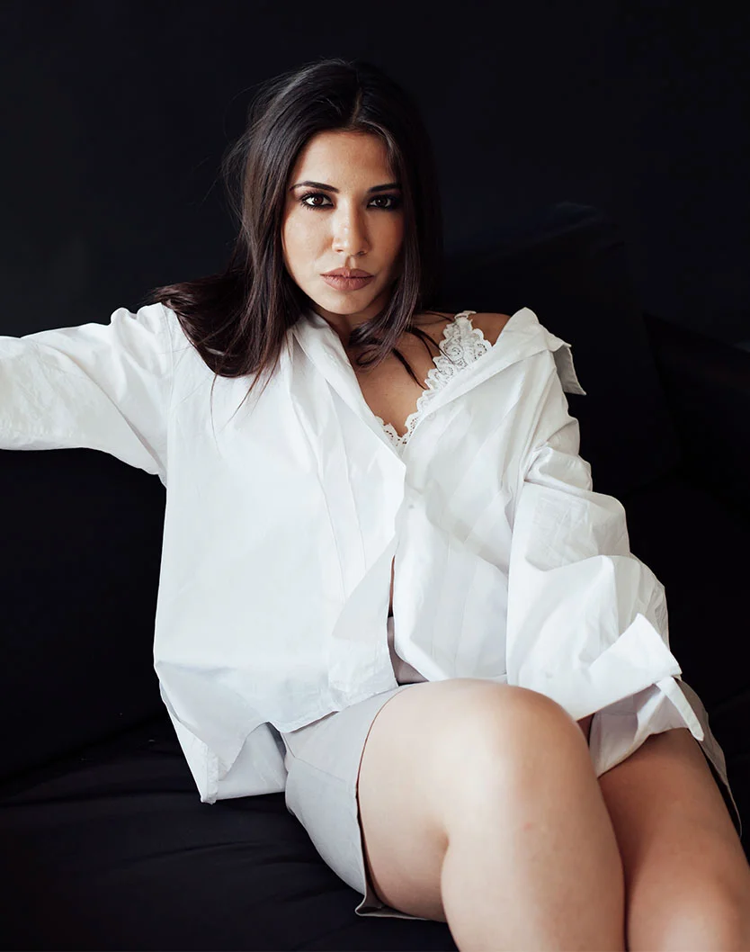 A woman with long, dark brown hair and light skin sits on a dark, likely black, couch or chair, her body angled toward the viewer in a relaxed posture. She wears an oversized white button-down shirt over a lacy white bralette-style top, with loose, flowy sleeves. Light taupe or gray shorts complete her outfit. Her expression is neutral to slightly serious, with visible makeup. Studio-style lighting highlights her skin tone and outfit against a solid black background, creating a striking contrast. - Dermal Fillers in Greenville, NC