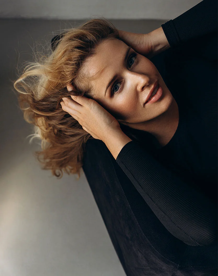 Close-up portrait of a woman with shoulder-length, light brown wavy hair that falls loosely around her face. She holds her head with both hands, her expression relaxed and slightly pensive. Her skin is light-toned with subtle makeup, and she is wearing a dark, long-sleeved pullover top. Soft, diffused lighting casts a gentle glow on her face and body. The background is a muted grayish-taupe, contributing to the calm, introspective mood of the image. - Facial Fat Grafting in Greenville, NC