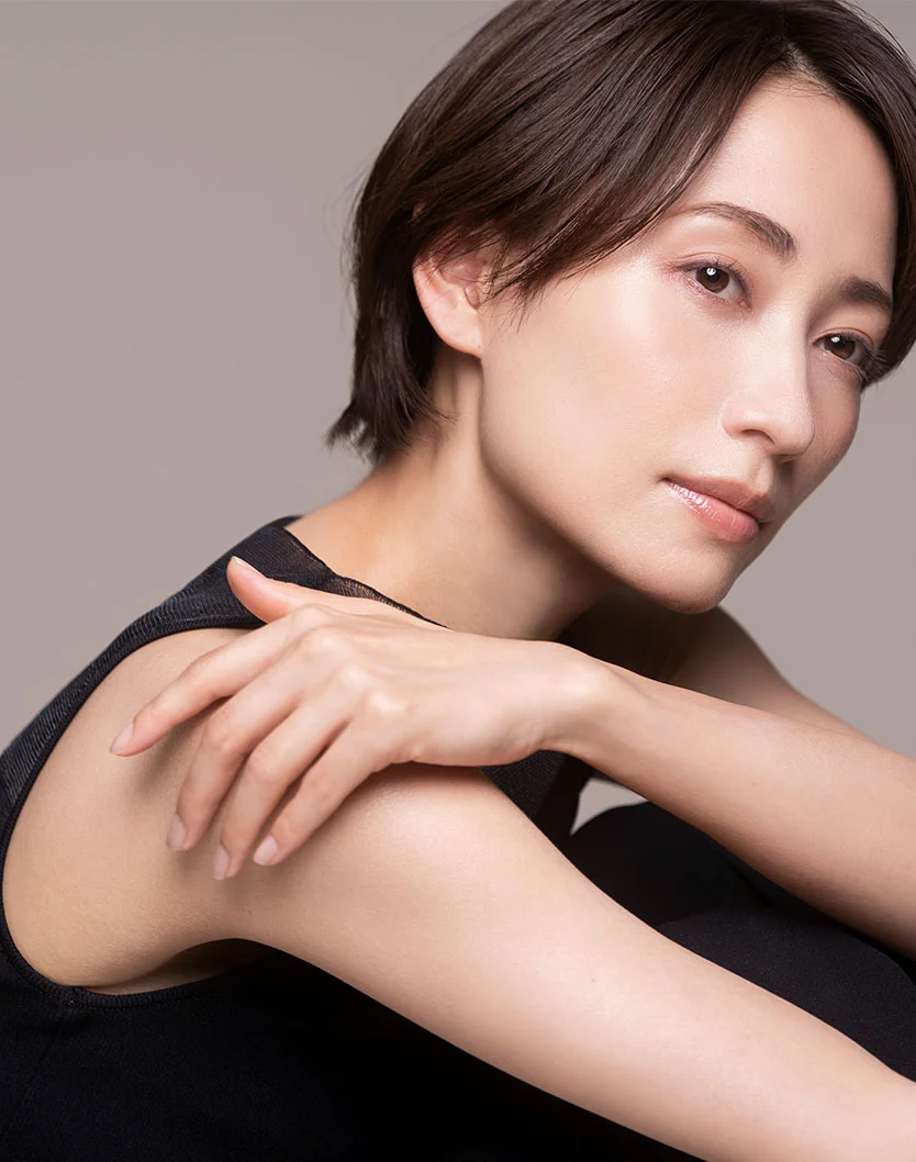 A seated woman with short dark brown hair and light skin, angled slightly toward the viewer. She wears a sleeveless black top with a sheer, silky texture. Her expression is neutral, arms resting softly on her torso. The background is muted gray-beige, with soft lighting highlighting her face and smooth skin. - Functional Hand Surgery in Greenville, NC
