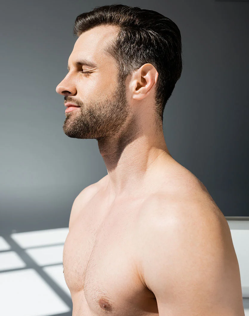 Close-up profile of a light-skinned man with short dark brown hair and a well-groomed beard. His eyes are closed, and he has a calm expression. He is shirtless, showing muscular chest and shoulders. The background is a neutral gray wall with soft shadows highlighting his skin and muscles. - Gynecomastia in Greenville, NC