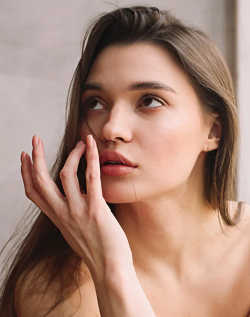 Close-up of a young woman with shoulder-length brown hair, gazing slightly upward and to the right with a thoughtful expression. Her hand rests near her face, and the soft lighting highlights her smooth, light-toned skin against a neutral gray background. - Lip Lift in Greenville, NC