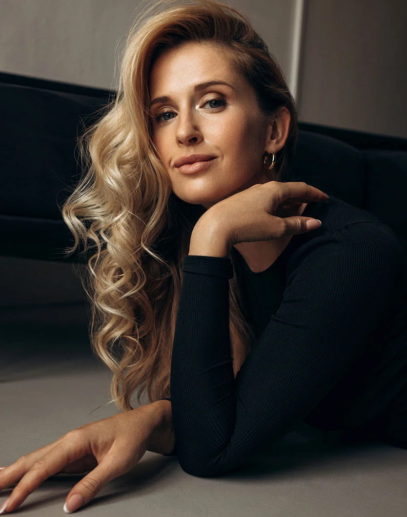 A woman with long, wavy blonde hair and smoky eye makeup reclines on a black leather couch. She wears a black jumper and supports her head with her left hand, showing a ring and French-manicured nails. Her blue eyes gaze directly at the camera. The image has a polished, professional look. - Lower Blepharoplasty in Greenville, NC