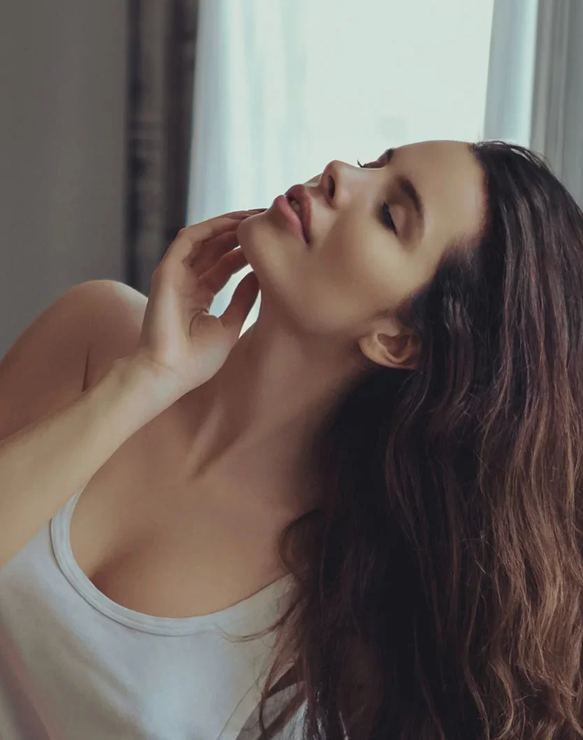 A portrait shot of a woman with dark long hair glancing up towards the ceiling. Her hand grazing against her neck, showing off comfort. She wears a white tank top and the background is blurred but a curtain is seen in the background. - Neck Liposuction in Greenville, NC